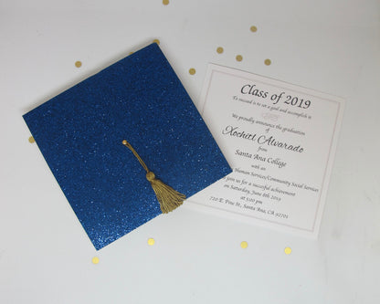 A blue glitter graduation cap invitation with a real gold tassel on a white background, accompanied by an envelope.