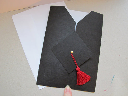 A folded black graduation invitation with a cap and gown design, featuring a red tassel and a gold brad,  with a white envelope.