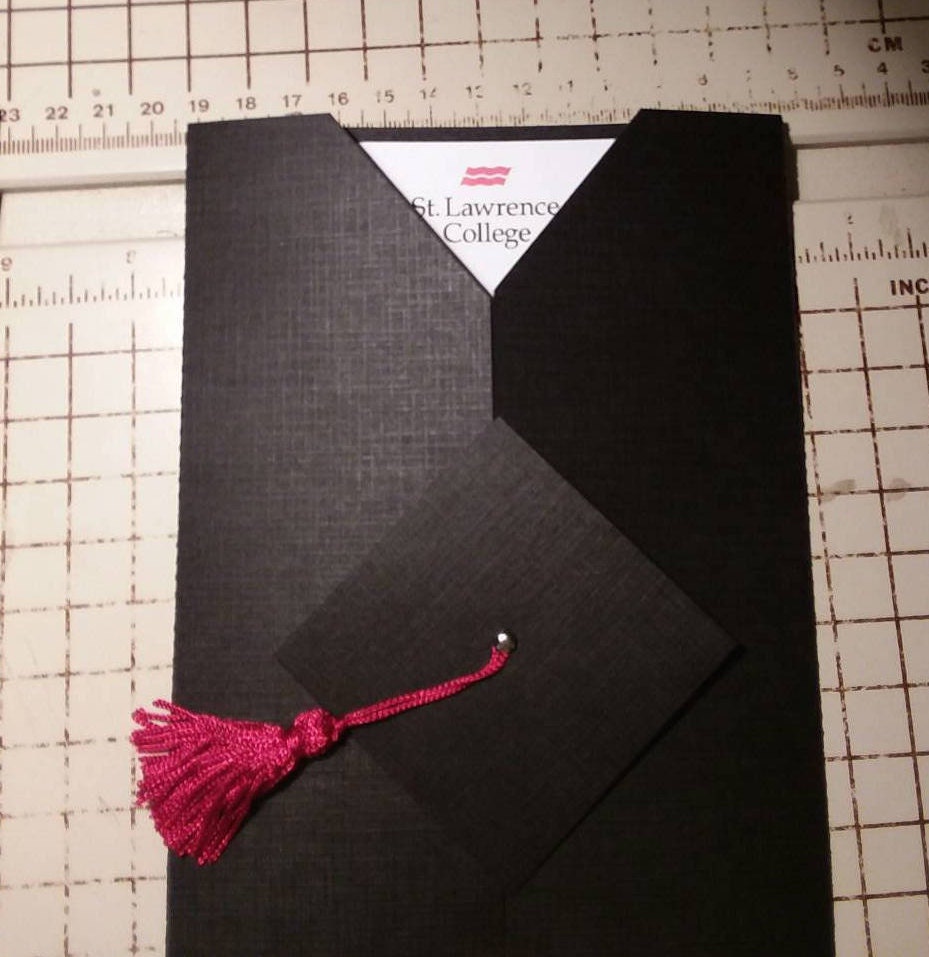 A folded black graduation invitation with a cap and gown design, featuring a red tassel and a gold brad and your college logo