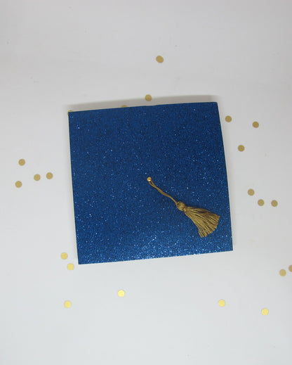 A blue glitter graduation cap invitation with a real gold tassel on a white background
