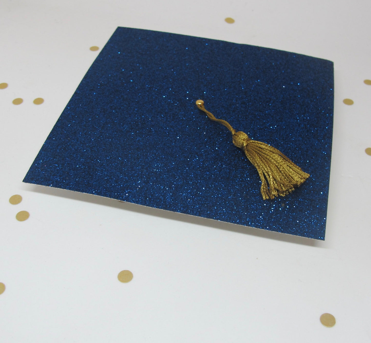 Up close view of a blue glitter graduation cap invitation with a real gold tassel on a white background